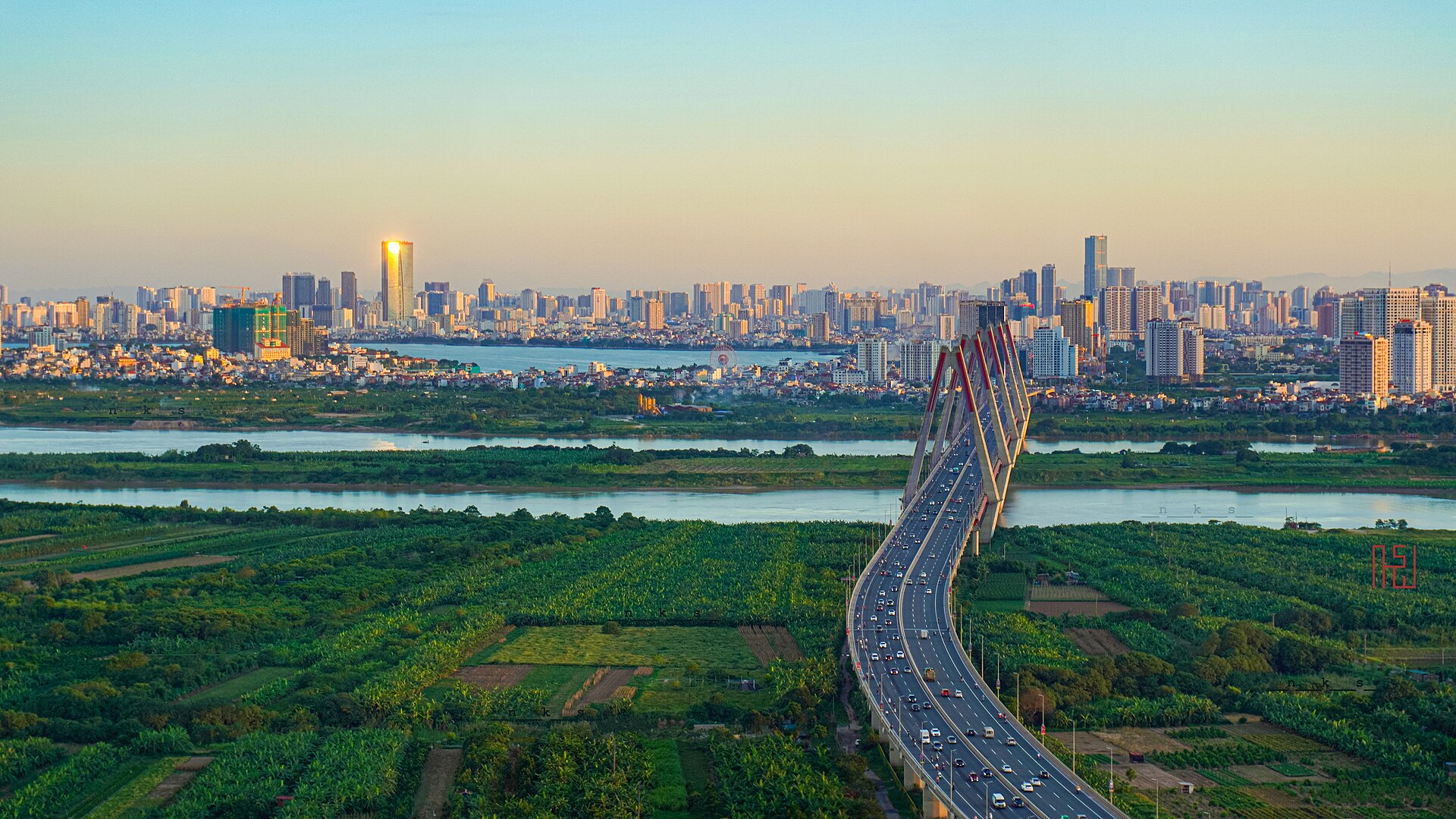 A bridge crosses the river toward the Hanoi skyline at sunset.